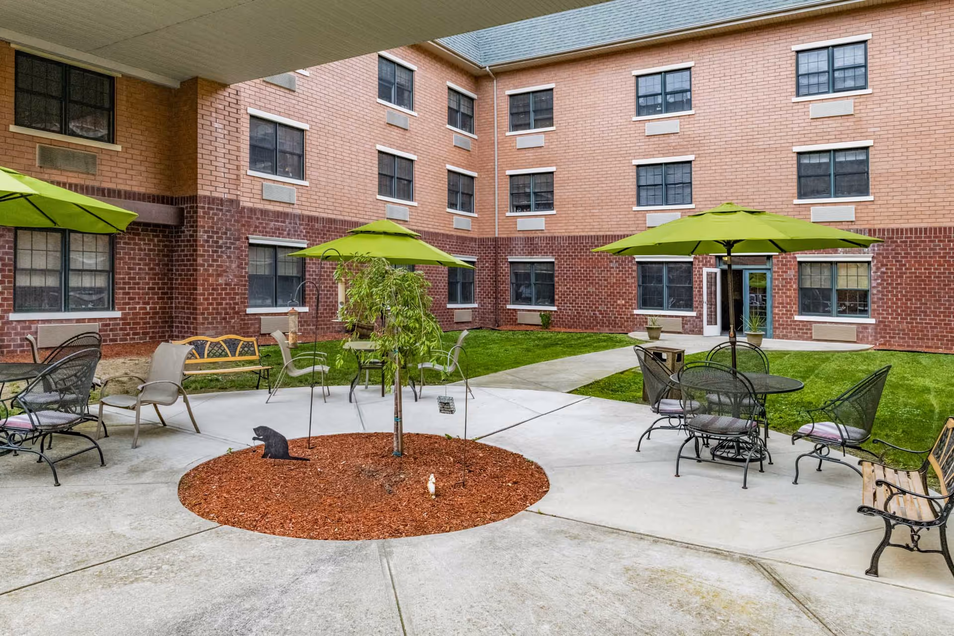 Brick three-story building surrounding a courtyard patio with tables, chairs, benches, and green umbrellas.
