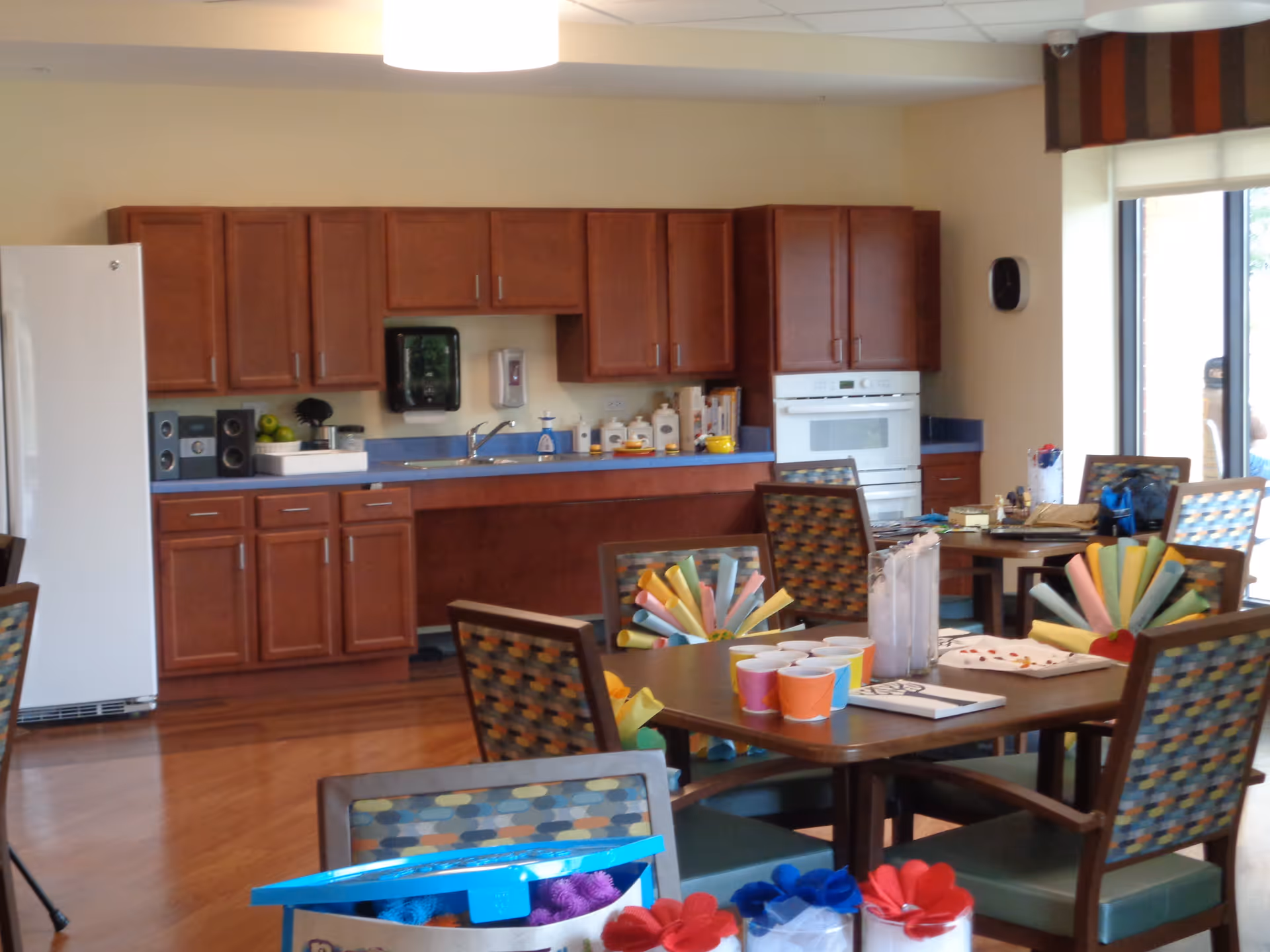 A kitchen and dining area in a supportive living community with wooden cabinets, a white refrigerator, a built-in oven, and a blue countertop. Several tables and chairs with patterned upholstery are arranged in the foreground, with colorful paper decorations and cups on the tables. Large windows allow natural light into the room.