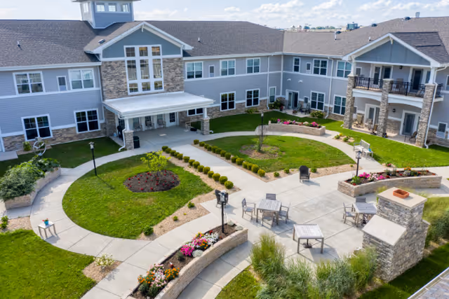 Outdoor courtyard area of a senior living facility with paved walkways, green lawns, flower beds, outdoor seating with tables and chairs, and a two-story building with balconies and large windows surrounding the courtyard.
