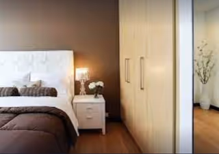 A modern bedroom featuring a white tufted headboard, a bed with brown bedding, a white nightstand with a lamp and a small vase of flowers, and a large wooden wardrobe with silver handles. A decorative plant is visible in the corner of the room.