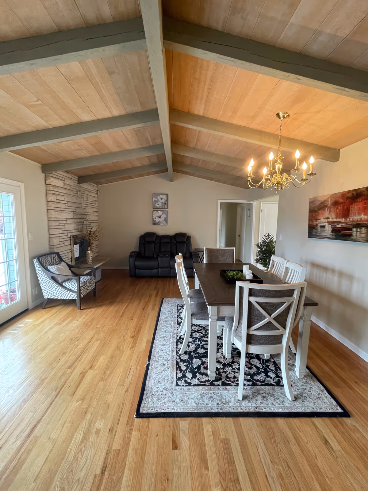 A spacious room with a wooden floor and a high ceiling with exposed beams. There is a dining table with six chairs on a patterned rug, a chandelier hanging above the table, a black leather recliner sofa against the far wall, a patterned armchair near a stone fireplace, and a large painting on the right wall.