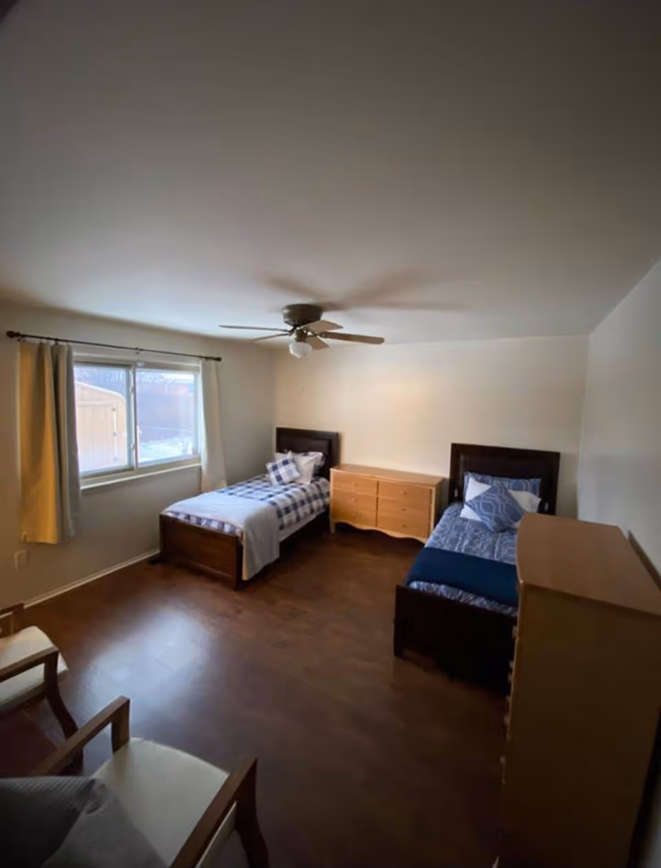 A bedroom with two twin beds, wooden dressers and chairs, a window with curtains, hardwood floor, and a ceiling fan.