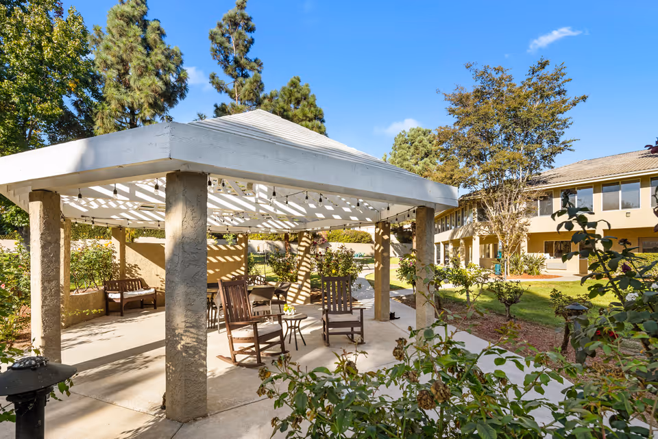 Covered gazebo with wooden chairs and small tables in a landscaped senior living courtyard with the facility building in the background.