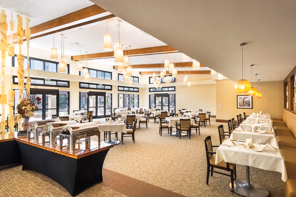 Spacious, bright dining room with multiple set tables, a buffet station, pendant lighting, and large windows.