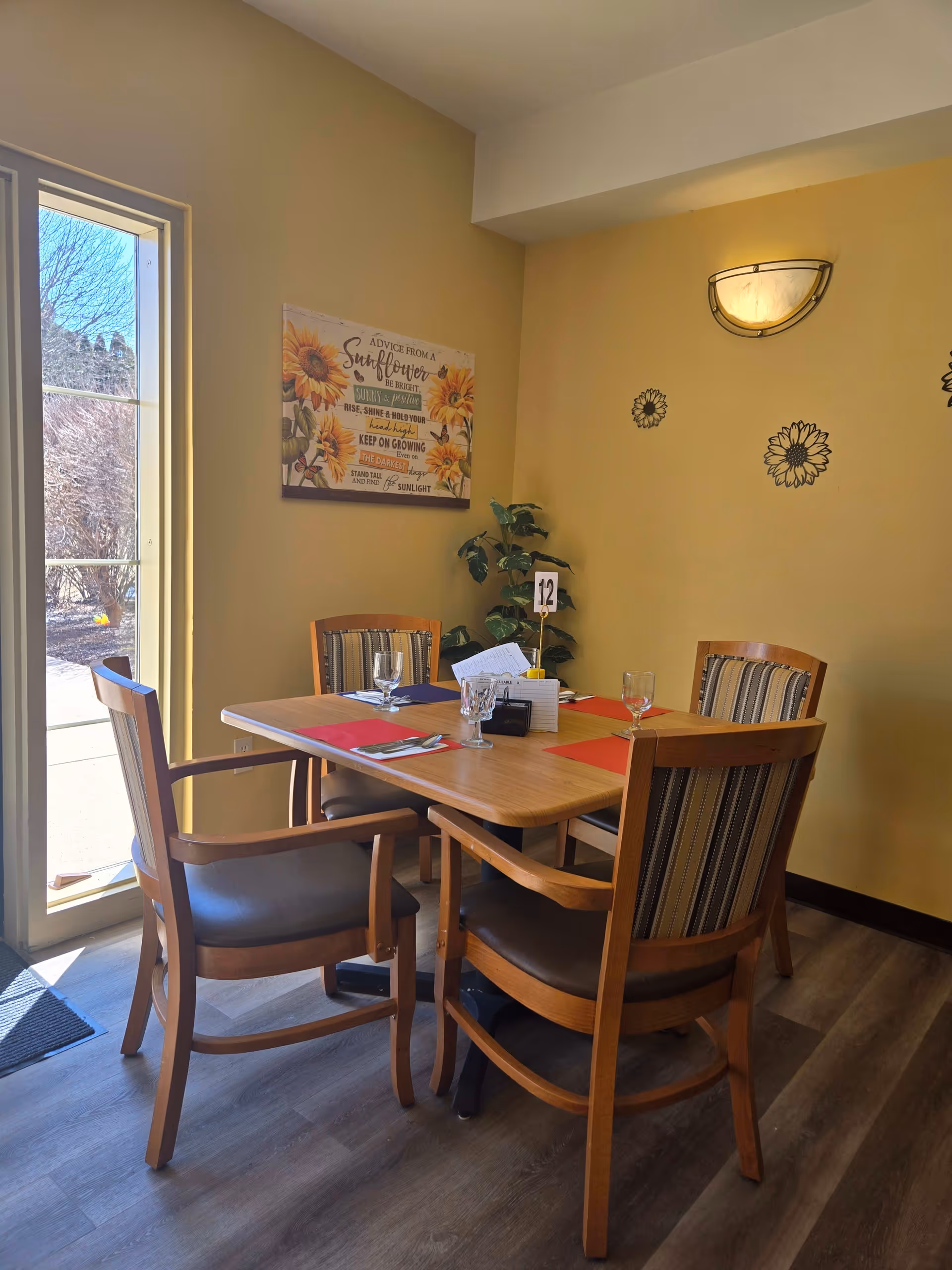 A dining area with a wooden table set for four, featuring four wooden chairs with striped cushions. The table has red placemats, glassware, and a small centerpiece with a table number 12. The room has yellow walls decorated with sunflower-themed wall art and a wall-mounted light fixture. A large window on the left side lets in natural light and shows an outdoor view with bare trees.
