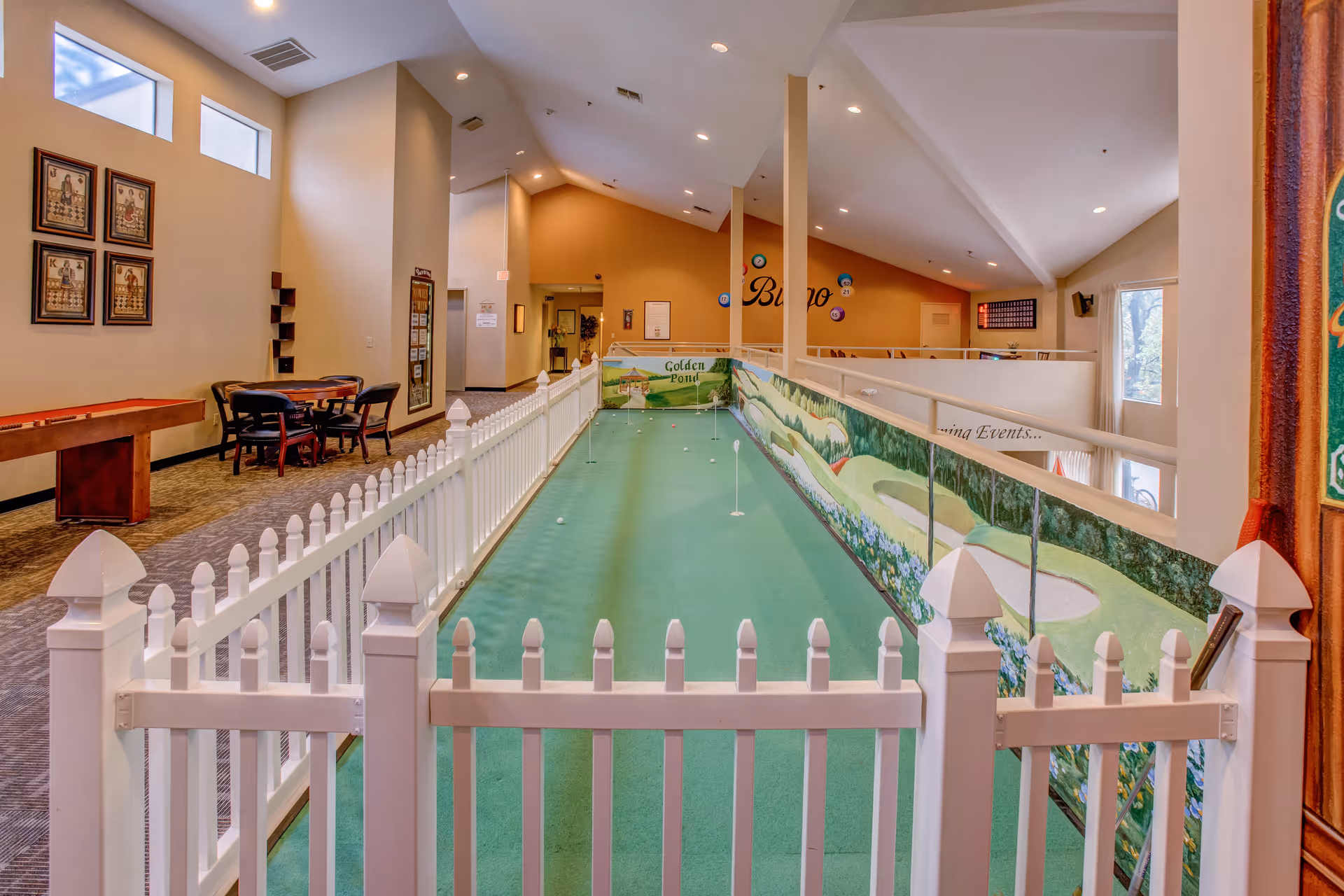 Indoor putting green area with a white picket fence surrounding it, located in a spacious room with high ceilings. The putting green has several golf balls and holes. On the left side, there is a shuffleboard table and a small table with four chairs. The walls are decorated with framed pictures and a mural of a golf course. The room is well-lit with recessed lighting and natural light from windows.