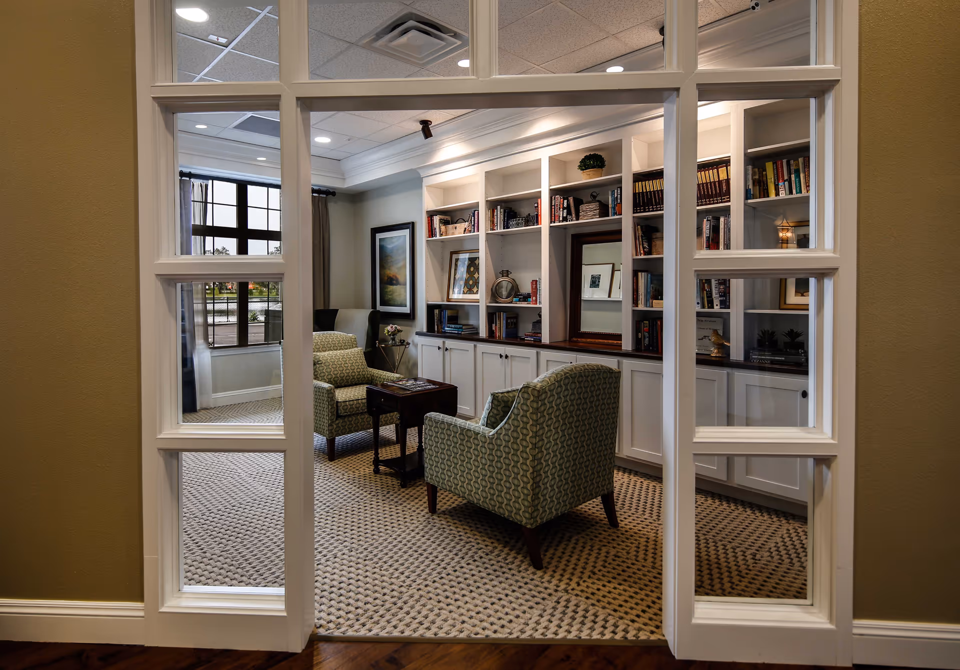 View through white-framed glass doors into a cozy sitting room with two patterned armchairs facing a small wooden table. The room features built-in white bookshelves filled with books and decorative items, a framed picture on the wall, and a large window with curtains letting in natural light.