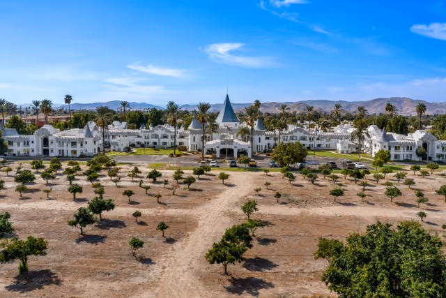Aerial view of Camelot - A Provincial Senior Living Community, featuring a large white castle-like building with multiple turrets and a central tower, surrounded by palm trees and a dry landscape with scattered small trees and bushes under a clear blue sky.