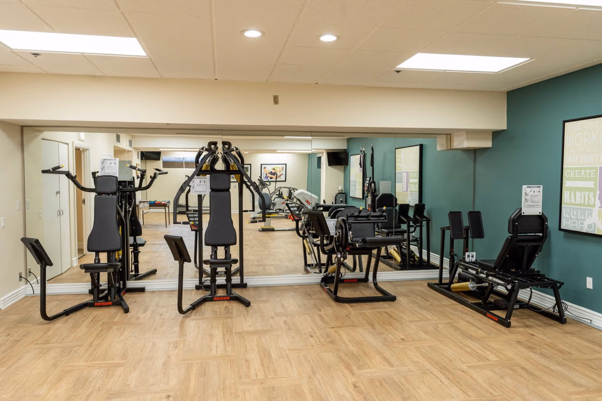 A fitness room with exercise equipment including weight machines and a leg press. The room has wood-patterned flooring, a large mirror covering one wall, teal painted walls, and motivational posters. The ceiling has recessed lighting.