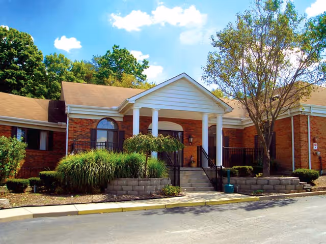 Front entrance of a single-story brick building with white columns, steps, shrubs, and trees under a blue sky.