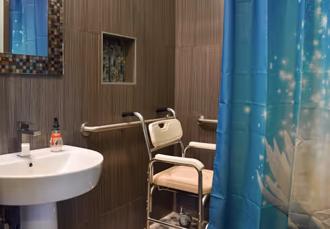 Accessible bathroom with a white pedestal sink, a wall-mounted mirror with a mosaic frame, a shower area with a blue curtain featuring a floral design, and a shower chair with armrests and wheels. The walls are covered with brown tiles and have grab bars installed for support.