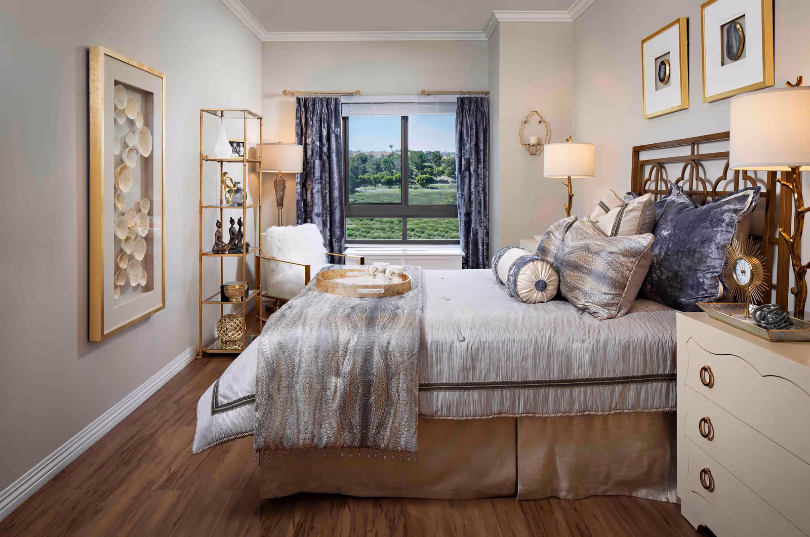 A well-decorated bedroom with a large bed covered in multiple pillows and a textured throw blanket. There is a window with dark curtains showing greenery outside. To the left of the bed is a gold metal shelving unit with decorative items and a white cushioned chair. On the right side is a cream-colored nightstand with a lamp and a decorative clock. The walls are light-colored with framed artwork and a wall-mounted candle holder.