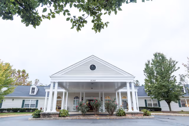 Front exterior view of a single-story building with white siding and a covered entrance supported by white columns. The building has a triangular pediment with a circular window and the number 10140 displayed. There are bushes and trees around the entrance and a paved driveway in front.