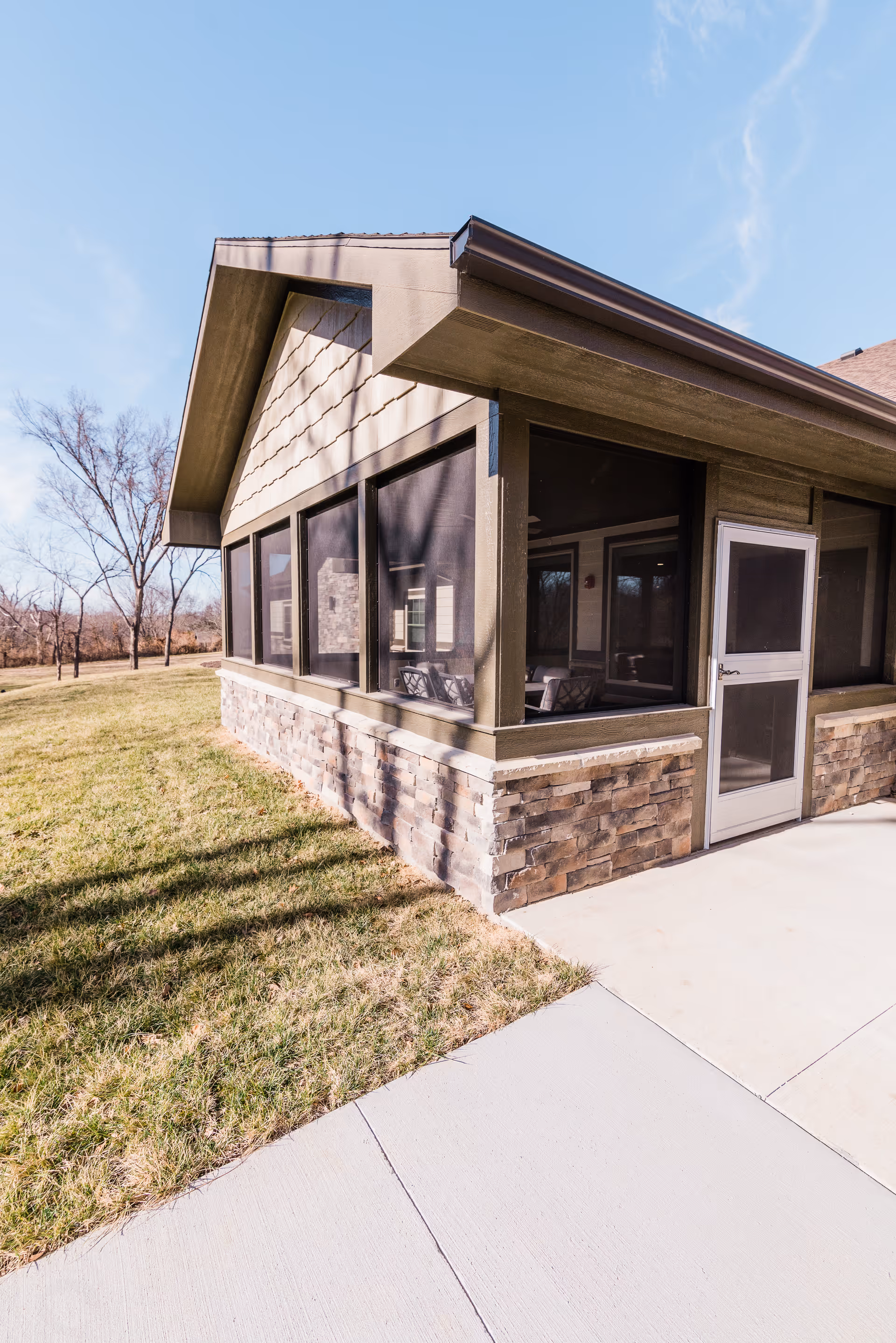 Screened porch of a cottage-style building with a stone veneer base, grassy lawn and concrete walkway under a clear blue sky.