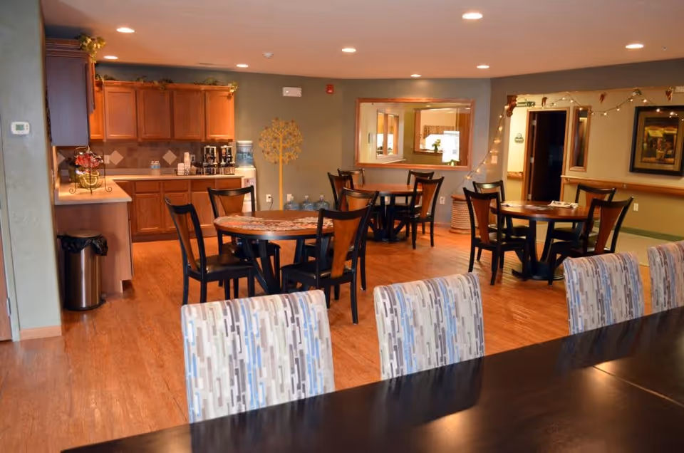 Community dining area with multiple round tables and chairs and a kitchenette in the background.