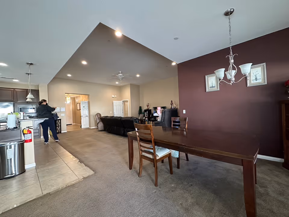 Interior view of a living and dining area in an assisted living facility. The foreground shows a wooden dining table with two chairs on a carpeted floor. The background features a living room with a dark sectional sofa, a TV, and a person standing near the kitchen area, which has dark cabinetry and stainless steel appliances. The walls are painted beige and dark brown, with framed artwork and a ceiling light fixture above the dining table.