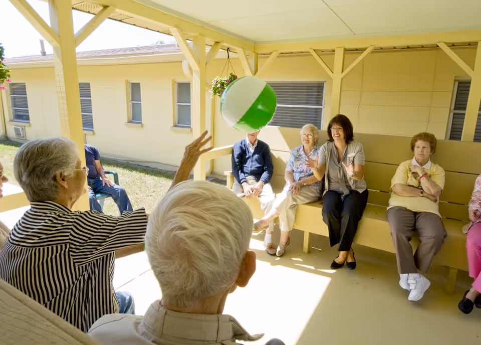 A group of elderly people sitting on benches under a covered outdoor patio, playing with a green and white beach ball. The setting is bright and sunny with a yellow building in the background.