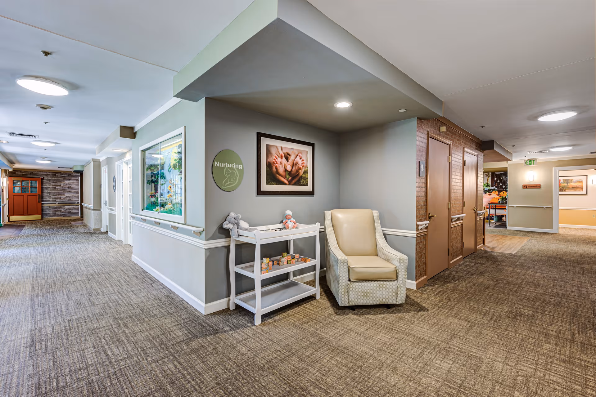 Interior hallway of a senior living facility with beige carpet and light gray walls. There is a beige armchair and a small white shelf holding a doll and some toys under a framed picture of adult hands holding baby feet. A green circular sign on the wall reads 'Nurturing'. The hallway extends in two directions with doors and handrails along the walls, and an exit sign is visible in the distance.