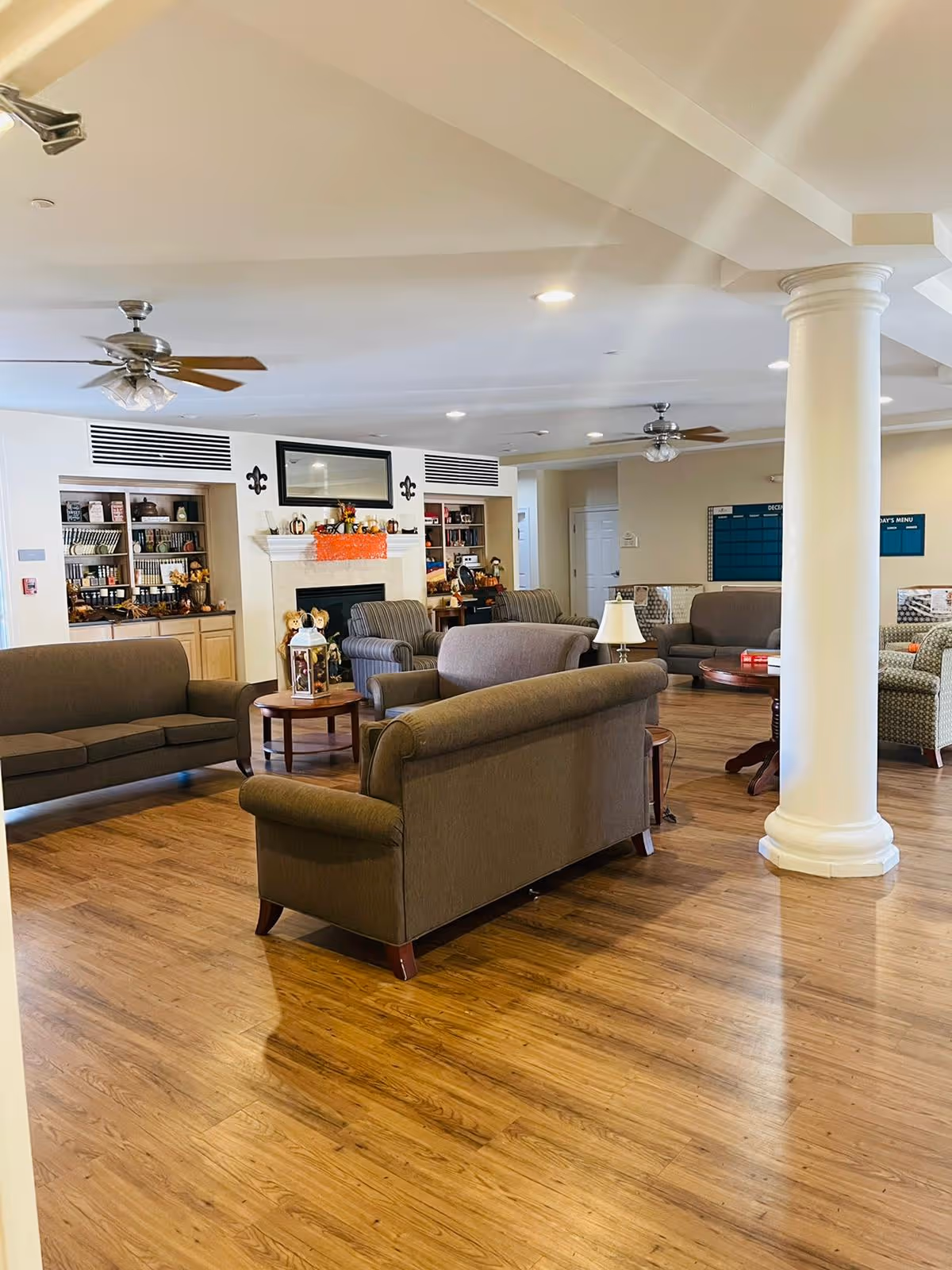 A spacious communal living room with multiple sofas and armchairs arranged around a fireplace and bookshelves, hardwood floors and a decorative column.