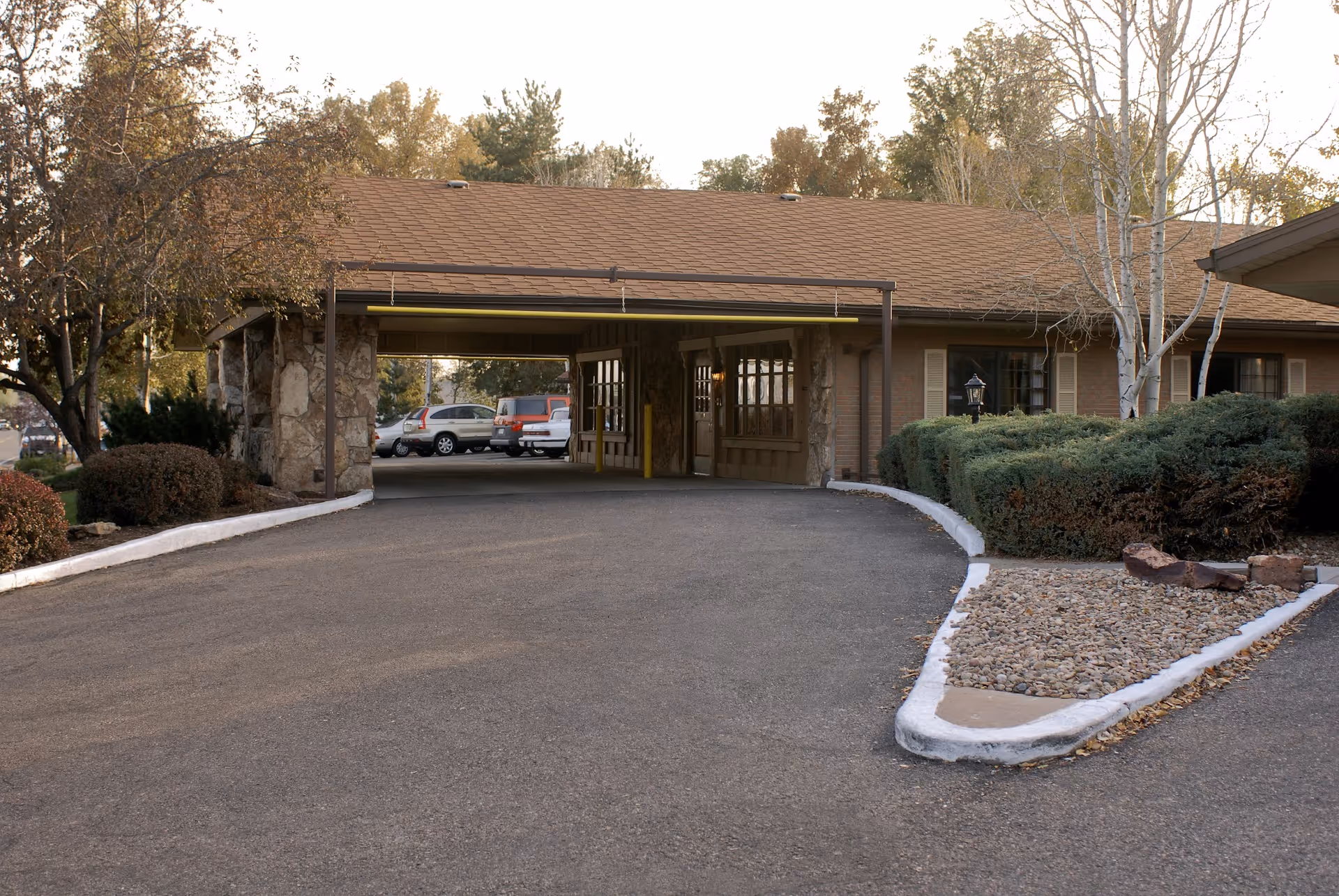 Exterior view of the entrance to Western Hills Health Care Center showing a covered driveway with stone pillars, parked cars in the background, and landscaped bushes and trees around the building.