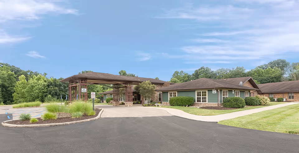 Single-story green and brick senior care building with a covered porte-cochère, driveway and landscaped grounds under a blue sky.