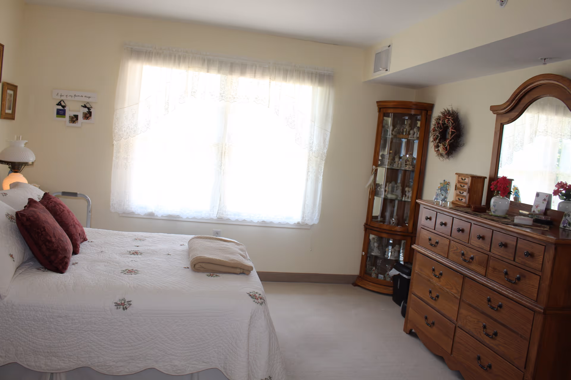 A bright bedroom with a large window covered by white lace curtains. The room features a bed with a white quilt and two red pillows, a folded beige towel on the bed, a wooden dresser with a mirror, and a glass display cabinet filled with decorative items. There is a wreath hanging on the wall above the dresser.