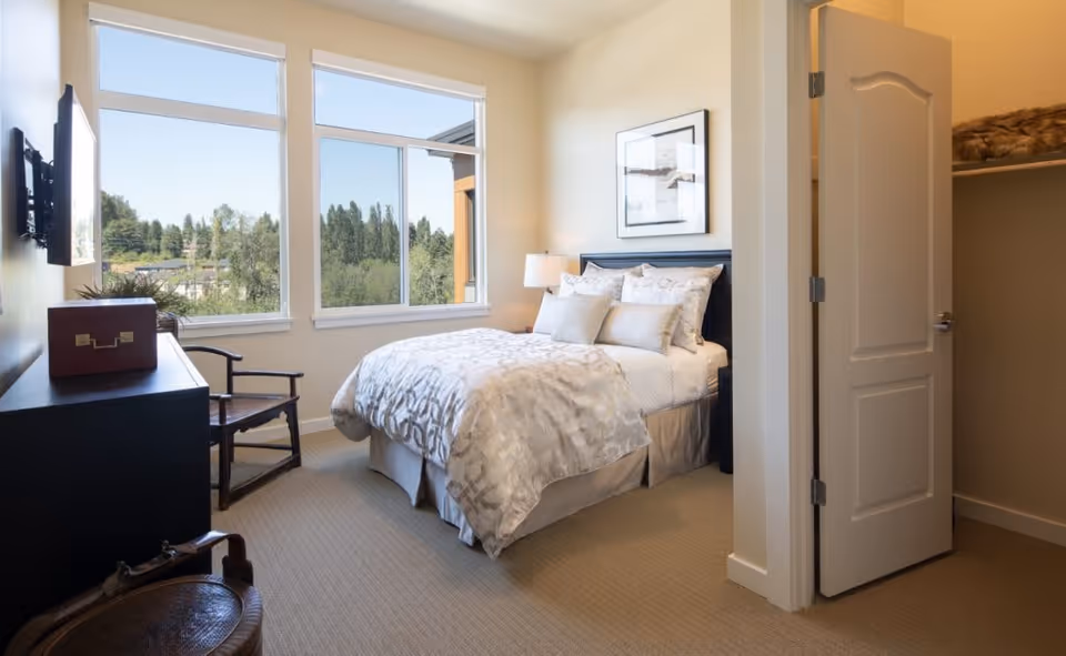 A bright bedroom with a neatly made bed featuring multiple pillows and a patterned comforter. The room has large windows showing a view of trees outside, a black dresser with a decorative box on top, a wooden chair, and an open closet door revealing shelves with folded blankets.