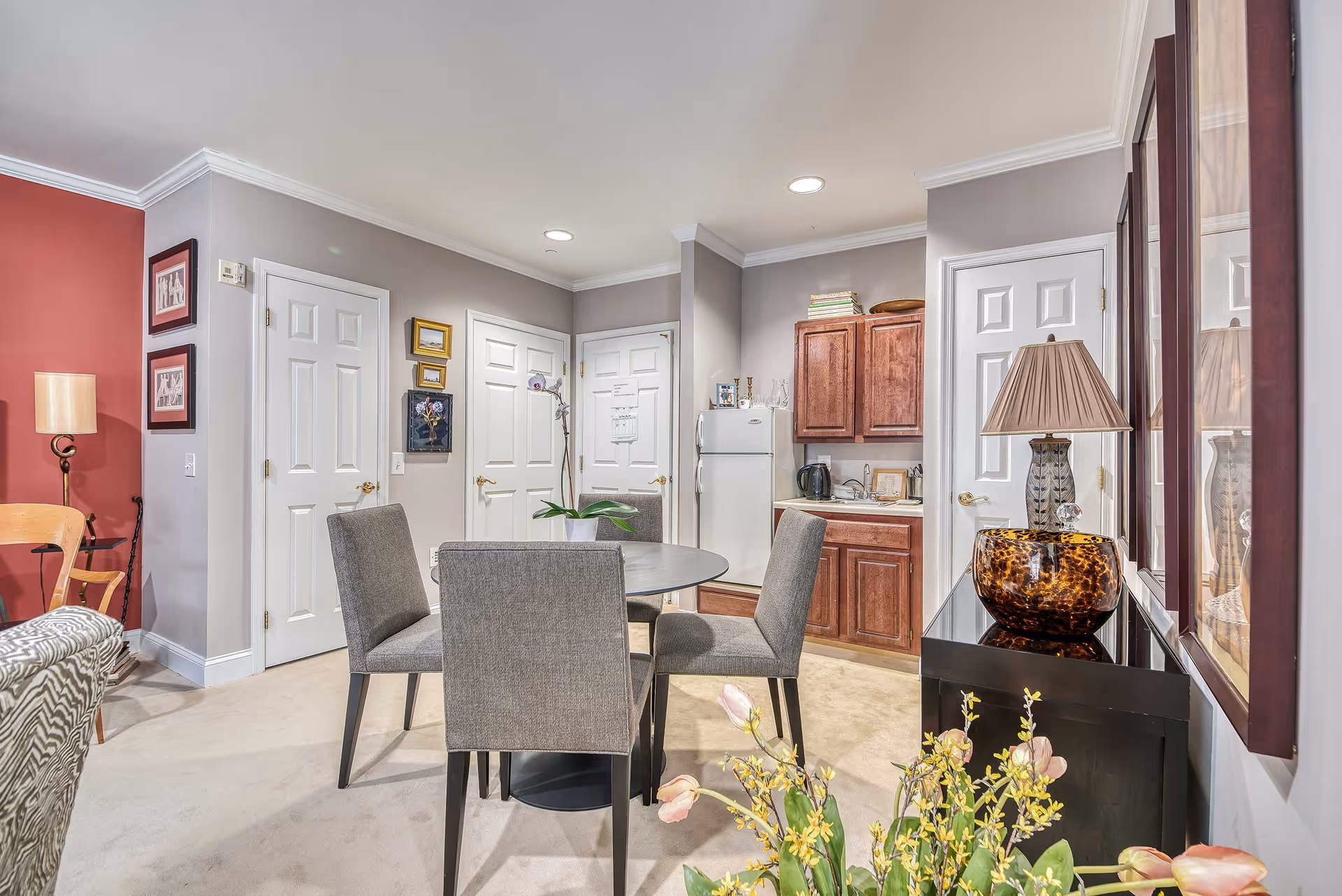 Round dining table with four gray upholstered chairs in a furnished apartment living area featuring a kitchenette, cabinets, and decorative lamps.