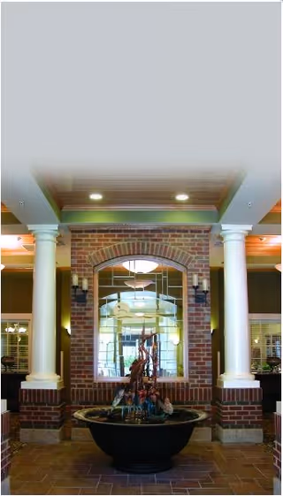 Interior view of a nursing and rehabilitation center featuring a decorative water fountain in a large black basin, positioned in front of a brick wall with an arched window. The space is framed by two white columns and illuminated by ceiling lights, with additional lighting fixtures on the brick wall.