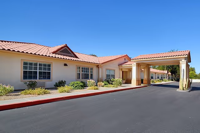 Single-story senior living building with a covered entrance, red tile roof, and landscaped shrubs under a clear blue sky.