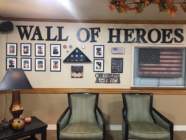 Two upholstered chairs and a side table beneath a 'WALL OF HEROES' display of framed photos and American flags on a wall.