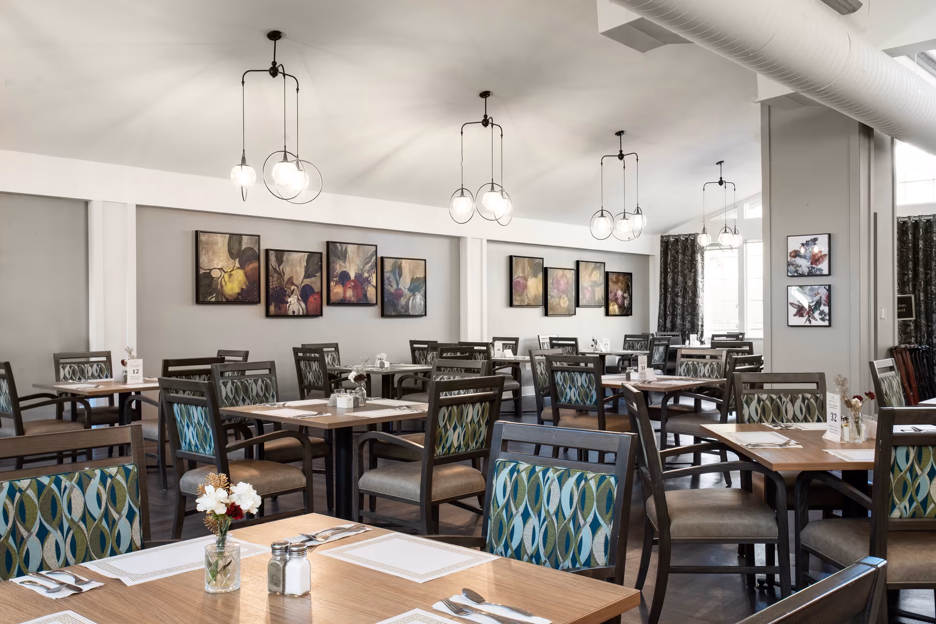 A spacious dining room with multiple wooden tables and chairs arranged neatly. Each table is set with placemats, cutlery, and small flower vases. The walls are decorated with framed artwork, and modern pendant lights hang from the ceiling. Large windows with curtains allow natural light to brighten the room.