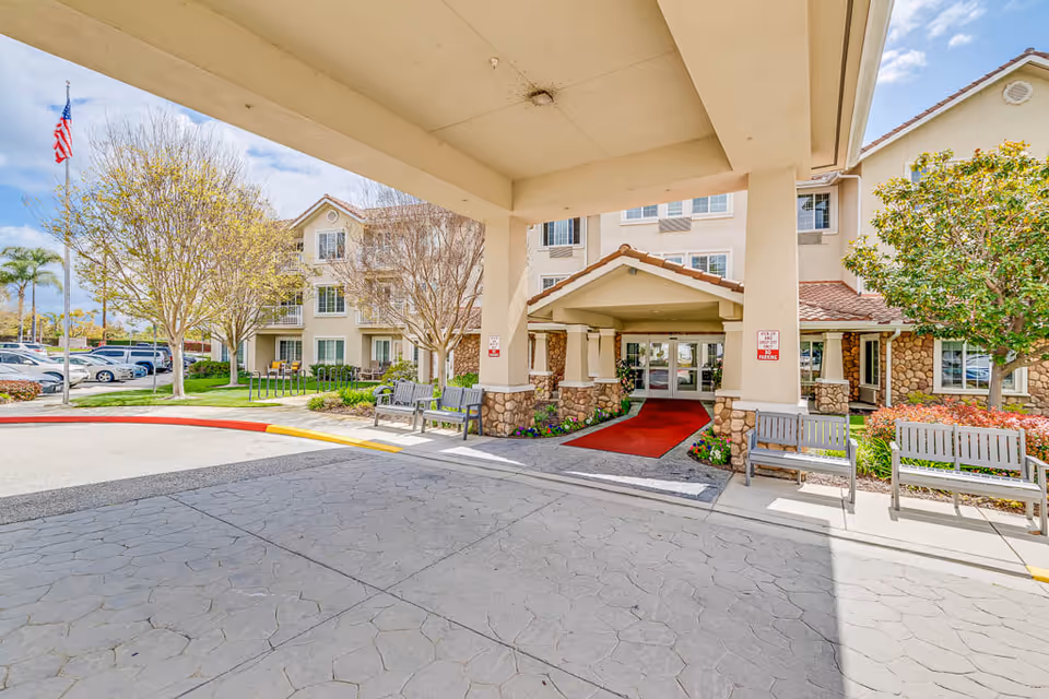 Entrance of The Palms At Bonaventure Assisted Living & Memory Care facility showing a covered drop-off area with benches on either side, a red carpet leading to glass doors, landscaped greenery, and a parking lot with cars and trees in the background under a partly cloudy sky.