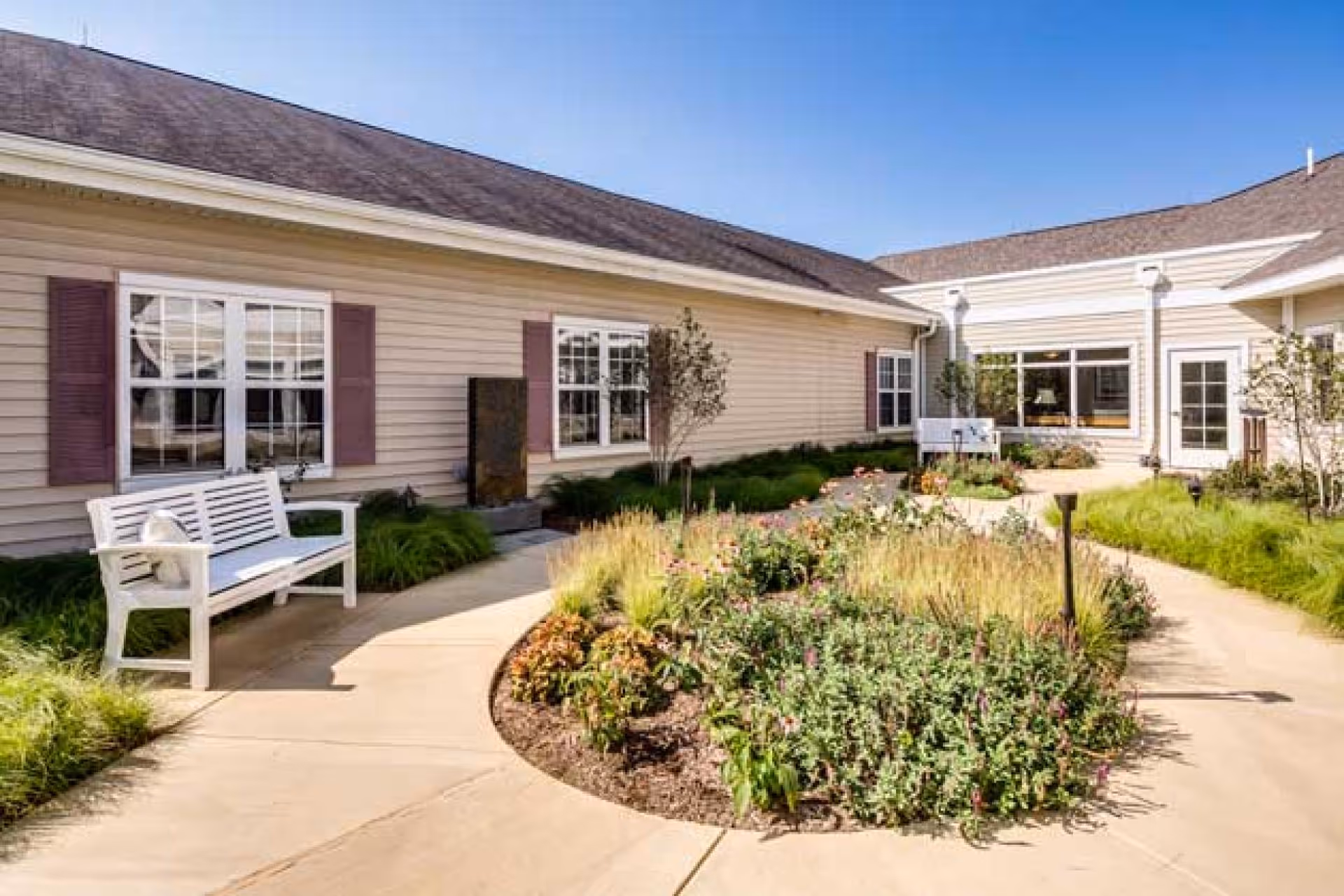 Sunny courtyard with a paved walkway, planted garden beds and white benches outside a single-story beige building.