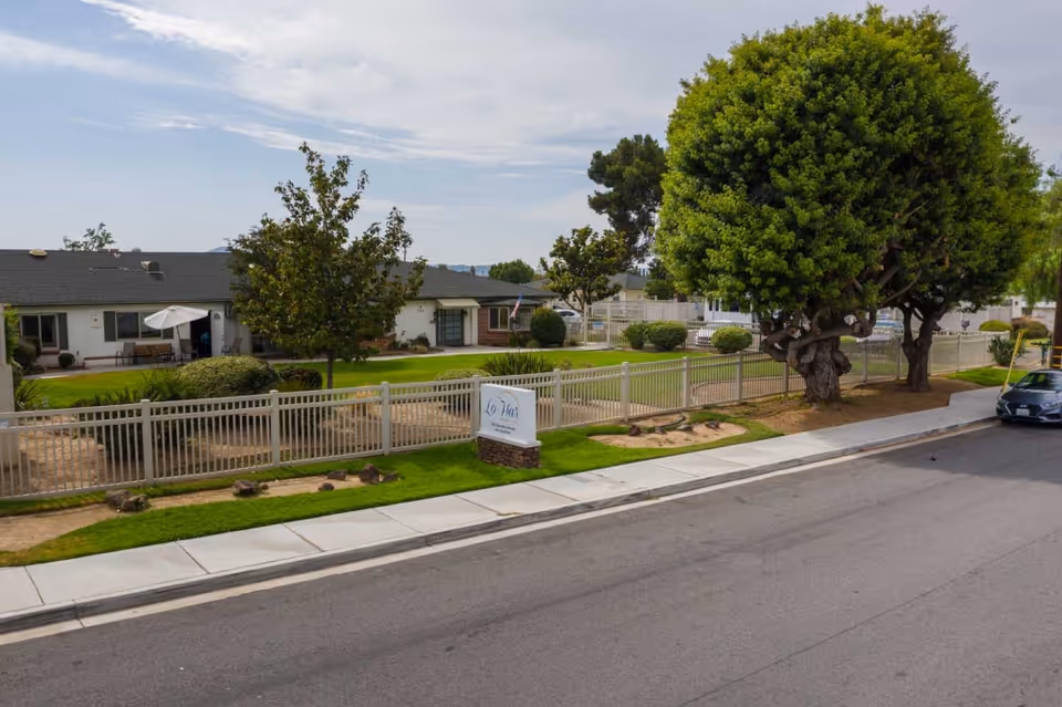 Street view of Lo-Har Senior Living facility showing a single-story building with a fenced green lawn, trees, and a sidewalk. A sign with the facility's name is visible near the sidewalk, and a car is parked on the street.