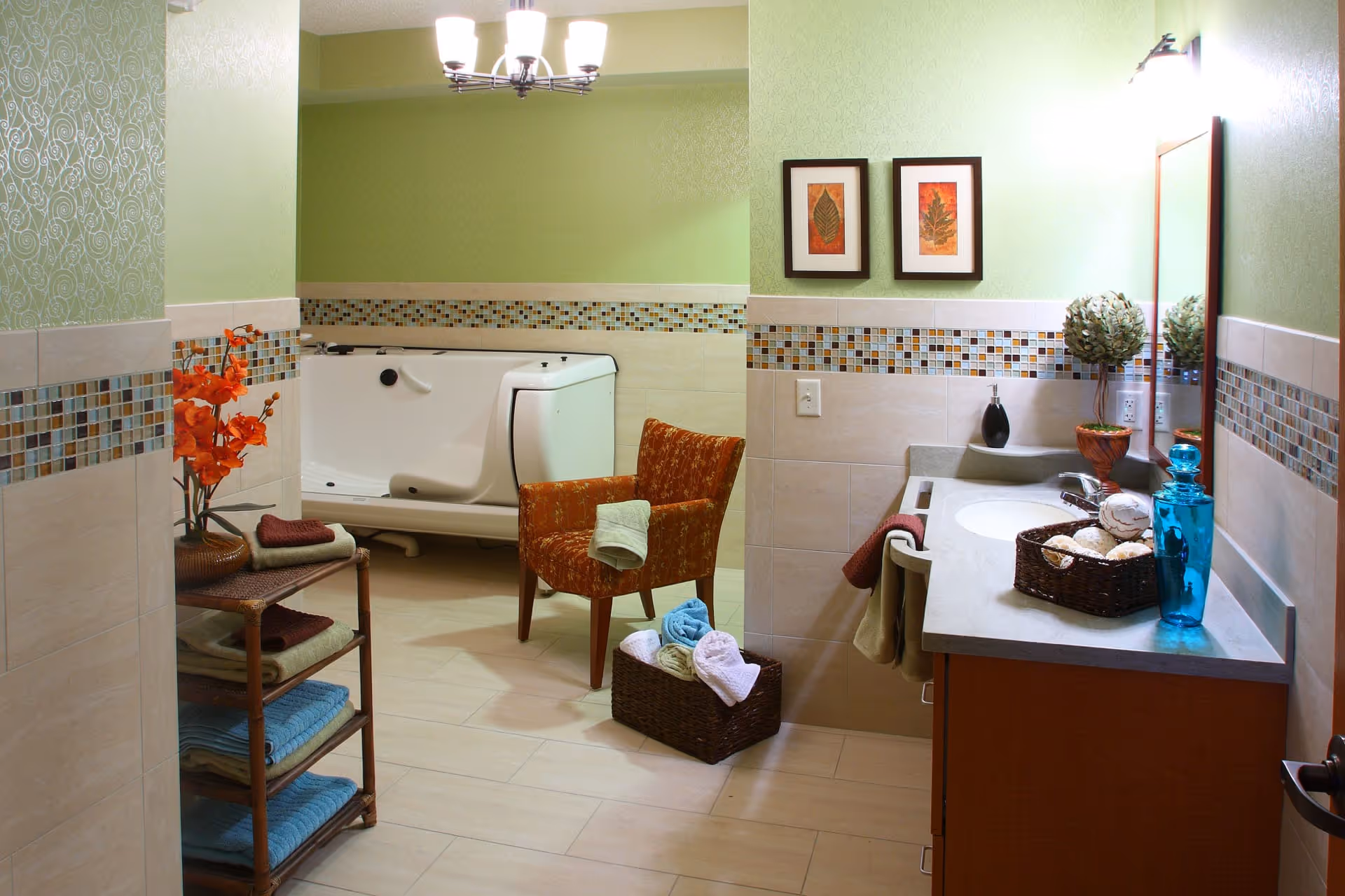 A spacious bathroom with light green walls and beige tiled floors. The room features a walk-in bathtub, an orange patterned armchair with a towel draped over it, a wooden shelving unit with folded towels, and a vanity with a sink, mirror, and decorative items including a blue glass bottle and a basket of decorative balls. Two framed leaf artworks hang on the wall above the bathtub.