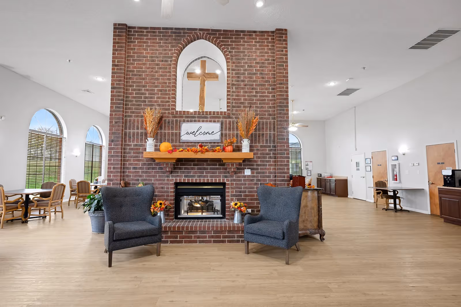 A spacious common area with a large brick fireplace in the center, decorated with autumn-themed items and a 'welcome' sign. Two gray armchairs are placed in front of the fireplace. The room has light wood flooring, white walls, and large arched windows letting in natural light. There are tables and chairs along the left side and a desk area on the right side of the room.