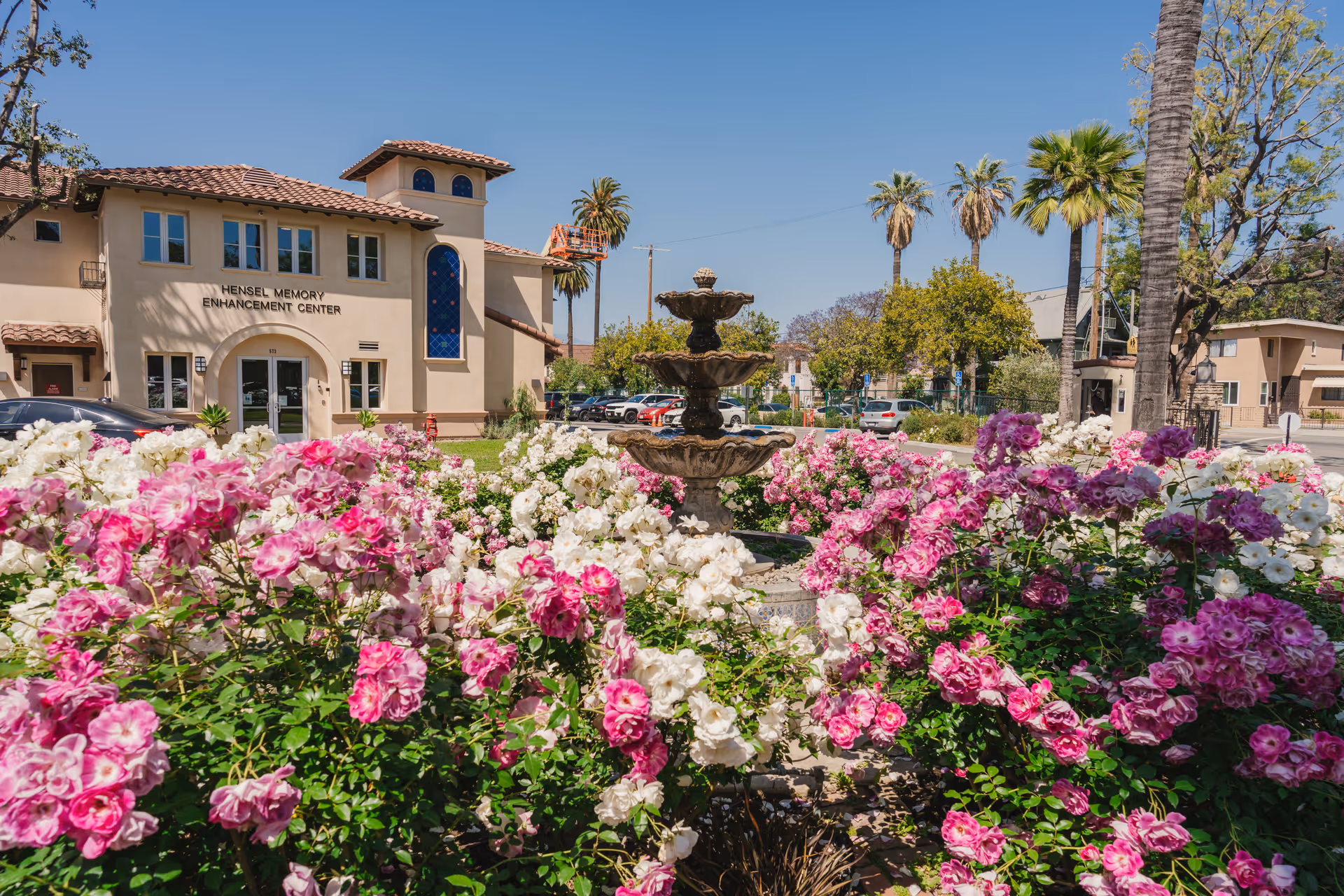 Outdoor view of the Hensel Memory Enhancement Center building at Hollenbeck Palms, with a garden full of pink and white flowers and a three-tiered stone fountain in the foreground. Palm trees and other greenery are visible under a clear blue sky.