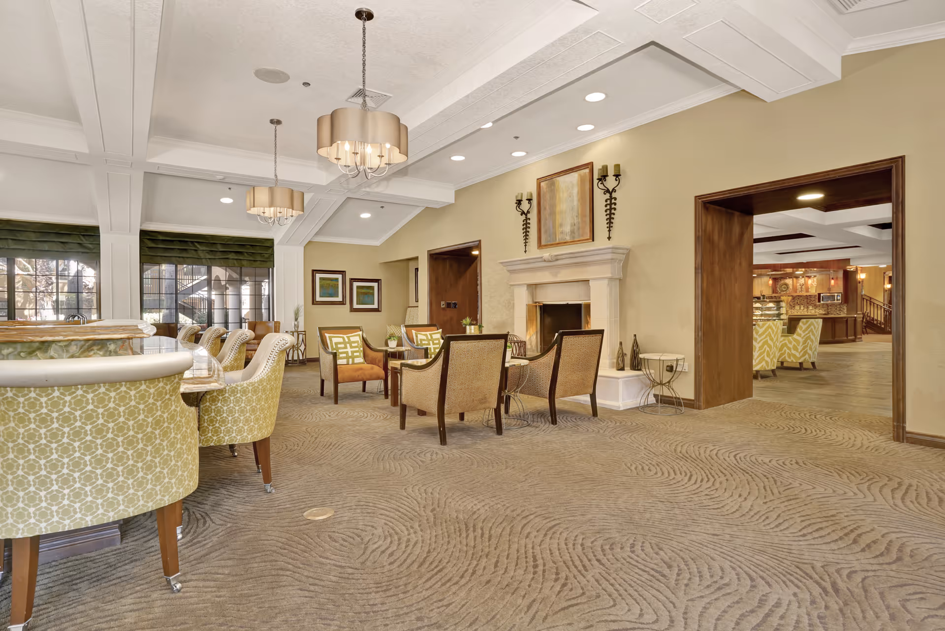 Bright communal lounge with patterned armchairs and tables, a fireplace, chandeliers, and an open view into an adjacent dining area.