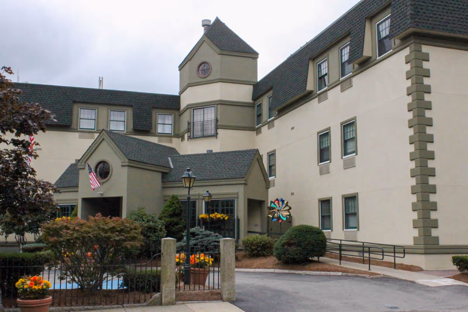 Exterior view of a multi-story senior living facility building with beige walls and green trim. The entrance features a small covered porch with an American flag, surrounded by landscaped bushes, flowers, and a decorative pinwheel. A paved driveway and wheelchair ramp are visible in front of the building under a cloudy sky.