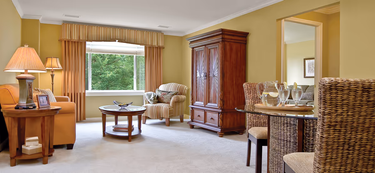 A cozy living room in a senior living community featuring a large window with beige curtains, a striped armchair with a decorative pillow, a wooden armoire, a round glass-top dining table with wicker chairs, a wooden coffee table, and a side table with a lamp and framed photo. The walls are painted light yellow and the floor is carpeted.