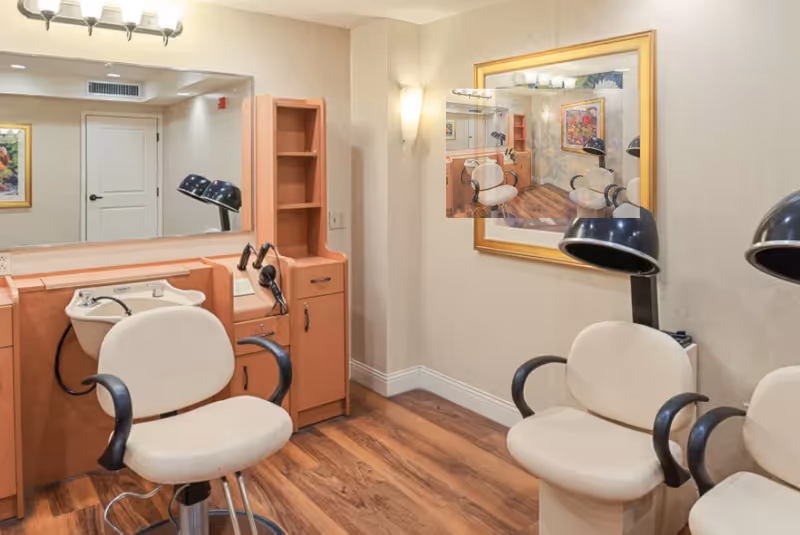Interior of a salon area with two beige salon chairs with black armrests, a hair washing station with a white sink, wooden cabinetry, a large mirror, and a framed floral artwork on the wall. The floor is wooden and the walls are light-colored.