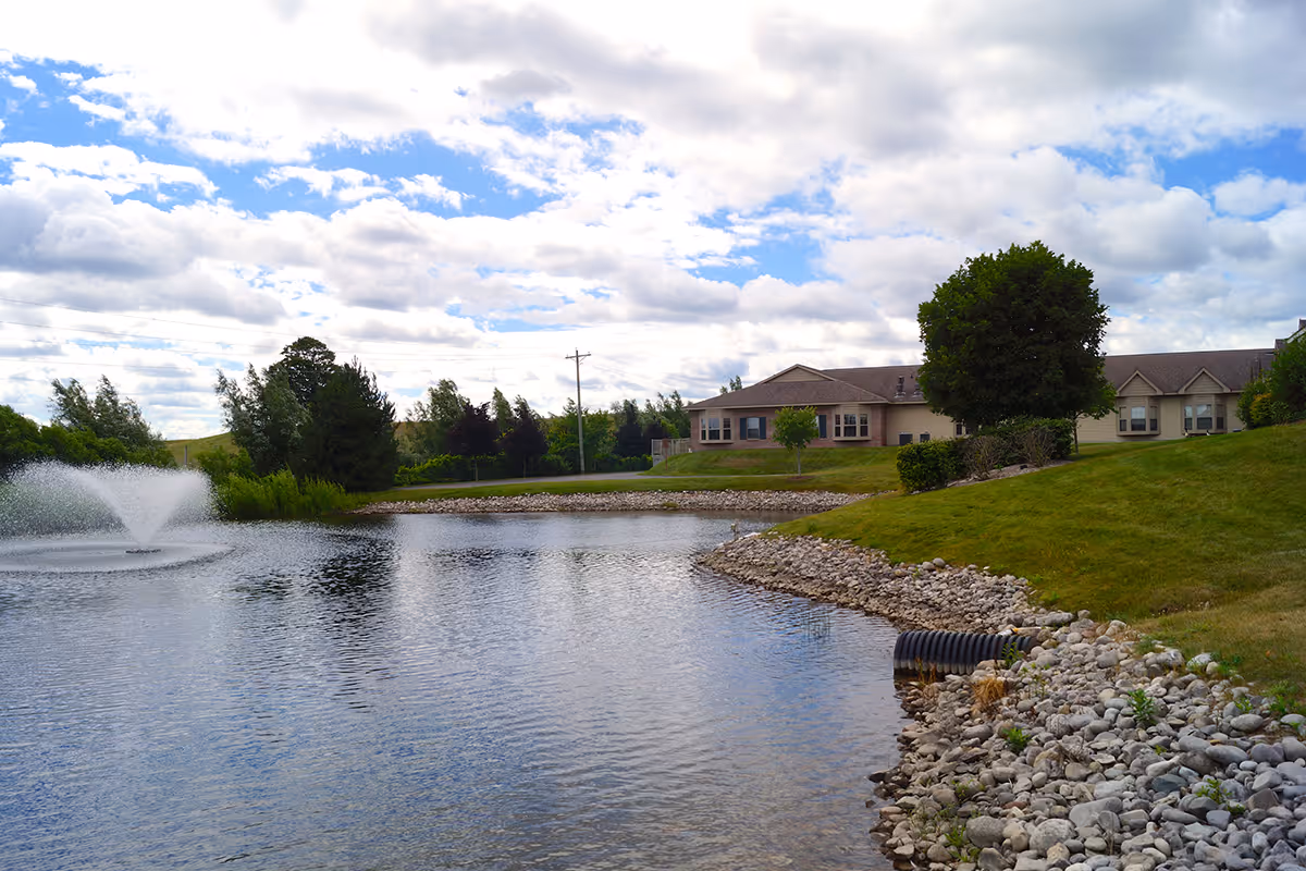 A serene outdoor scene at Bay Ridge Assisted Living Center featuring a pond with a water fountain, surrounded by green grass, trees, and a rocky shoreline. In the background, there are single-story buildings under a partly cloudy sky.
