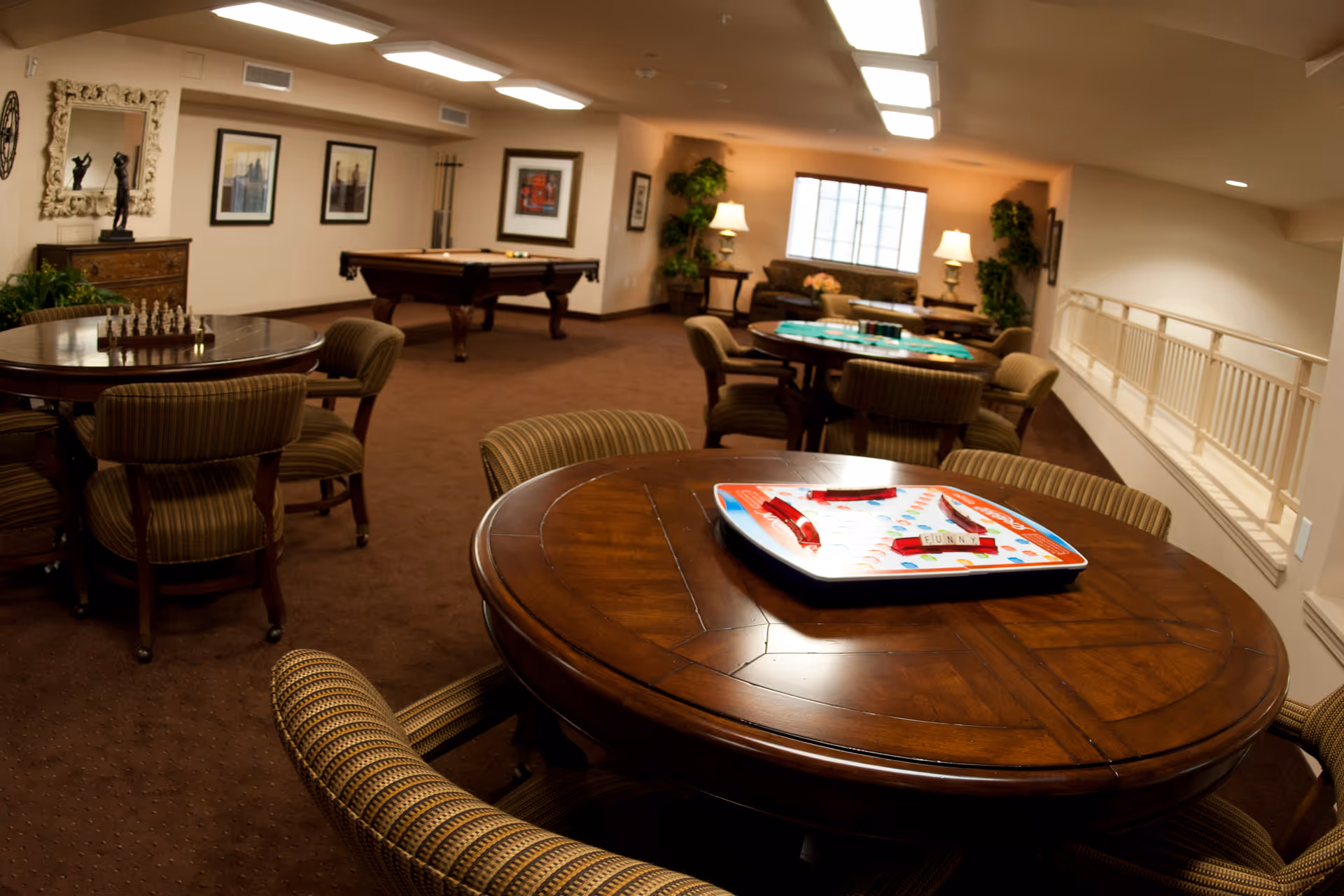 A cozy game room with several round wooden tables surrounded by cushioned chairs. One table has a chess set, another has a board game with tiles. In the background, there is a pool table and a seating area with sofas, lamps, and plants near a window.
