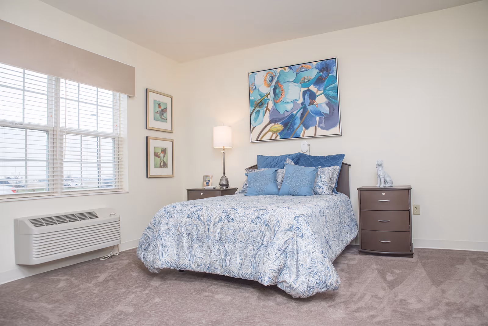 A bedroom with a bed covered in a blue and white patterned comforter and multiple blue pillows. There are two dark wood nightstands on either side of the bed, one with a lamp and picture frame, the other with a small white dog figurine. Above the bed hangs a large colorful floral painting. Two framed pictures are on the wall next to a window with white blinds and a beige valance. The floor is carpeted in a light brown color.