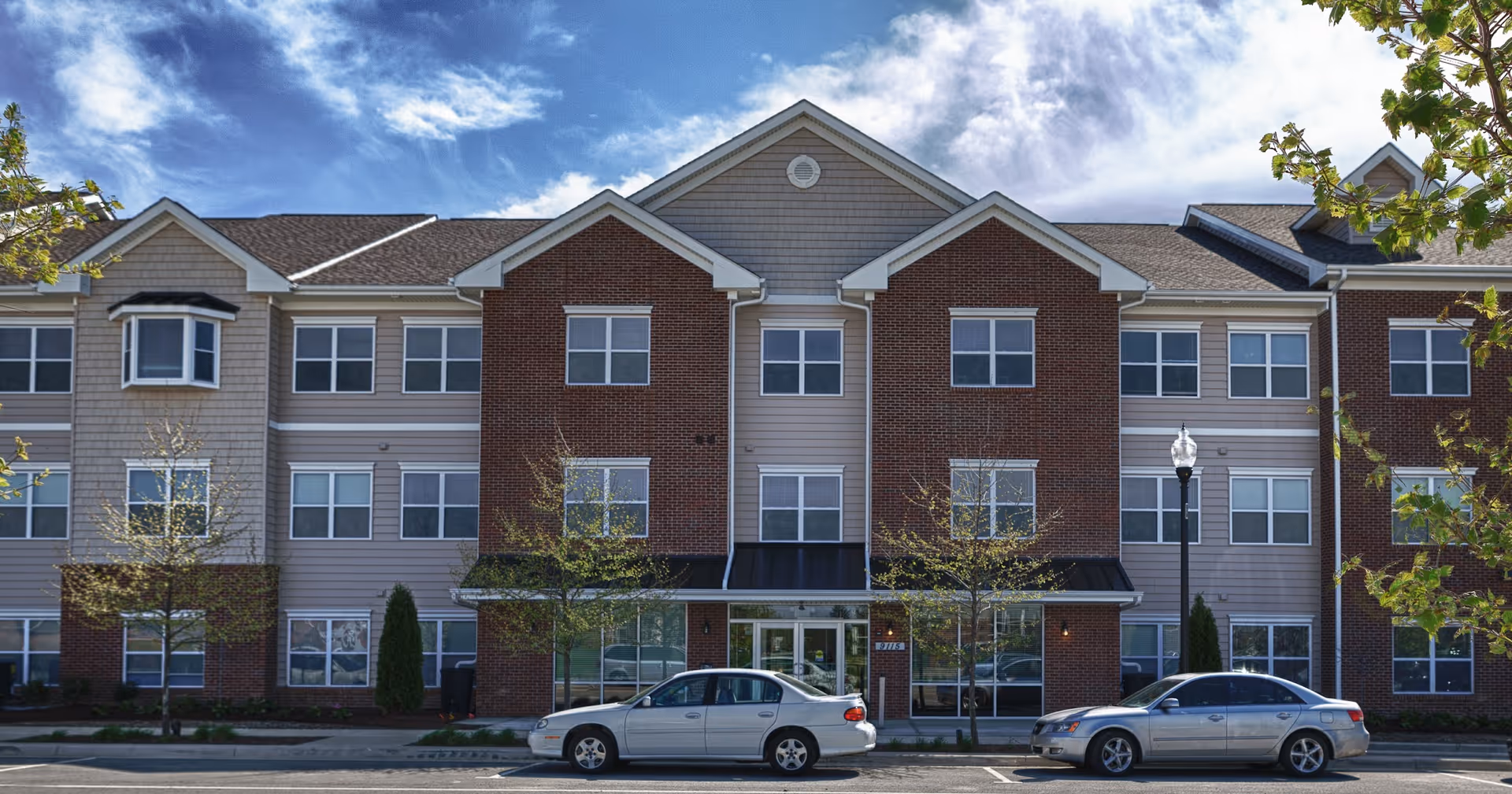 Front exterior view of a three-story senior living facility building with a combination of brick and siding facade, multiple windows, a central entrance with glass doors, two parked cars in front, and a partly cloudy sky above.