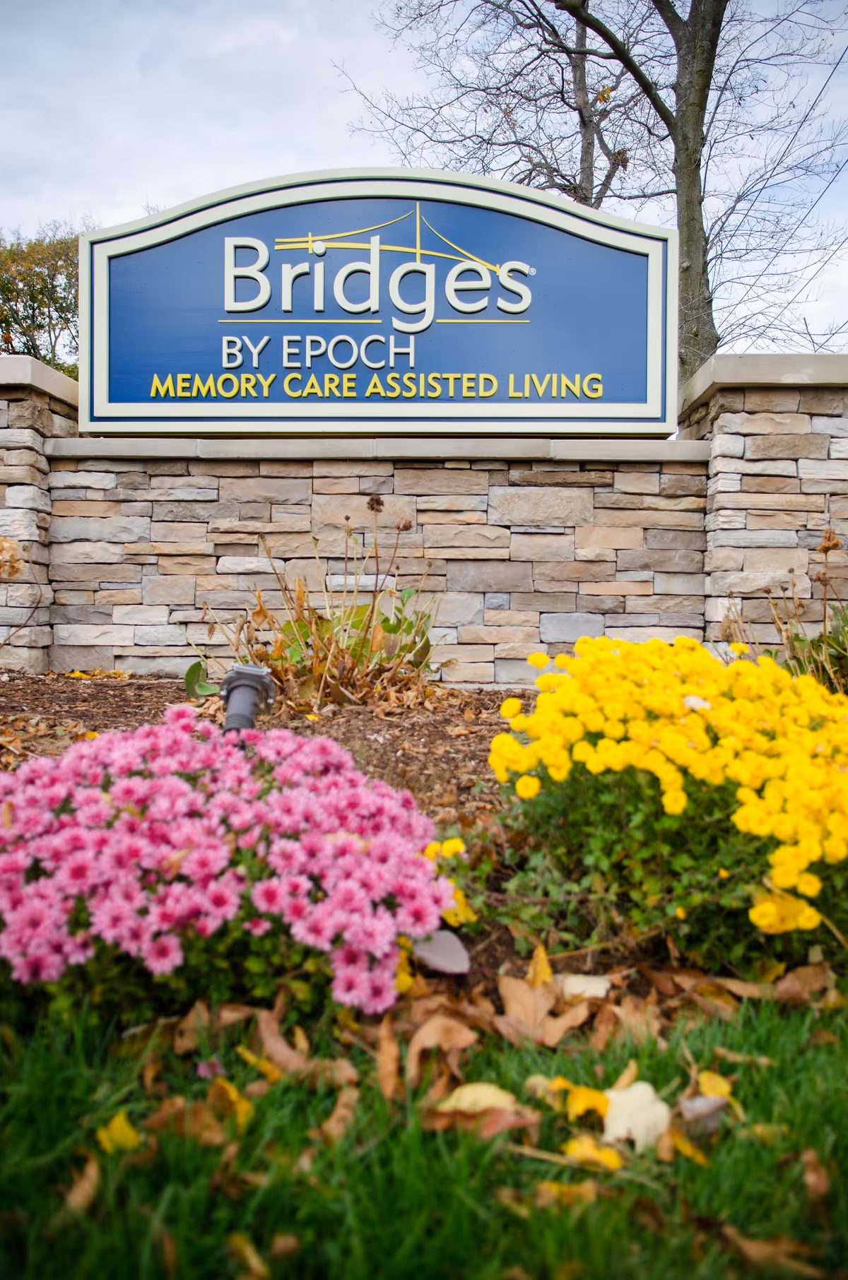 Stone sign for Bridges by EPOCH Memory Care Assisted Living with pink and yellow flowers in front and trees in the background.