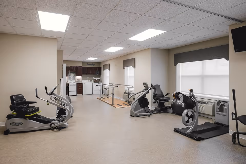 A well-lit exercise room in a senior living facility featuring various fitness equipment including recumbent bikes, an elliptical machine, and a treadmill. The room has beige walls, large windows with gray valances, and a ceiling with recessed lighting. In the background, there is a laundry area with a washer, dryer, and cabinets.