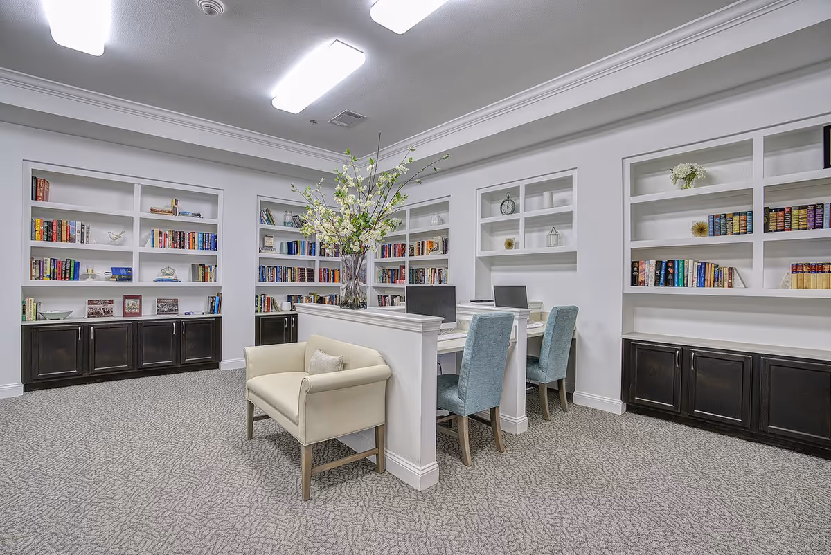 A bright and modern library or study area with built-in white bookshelves filled with books and decorative items. There are two computer workstations with blue upholstered chairs and a cream-colored armchair in the foreground. A large vase with white flowers is placed on the divider between the workstations. The room has a patterned carpet and overhead fluorescent lighting.