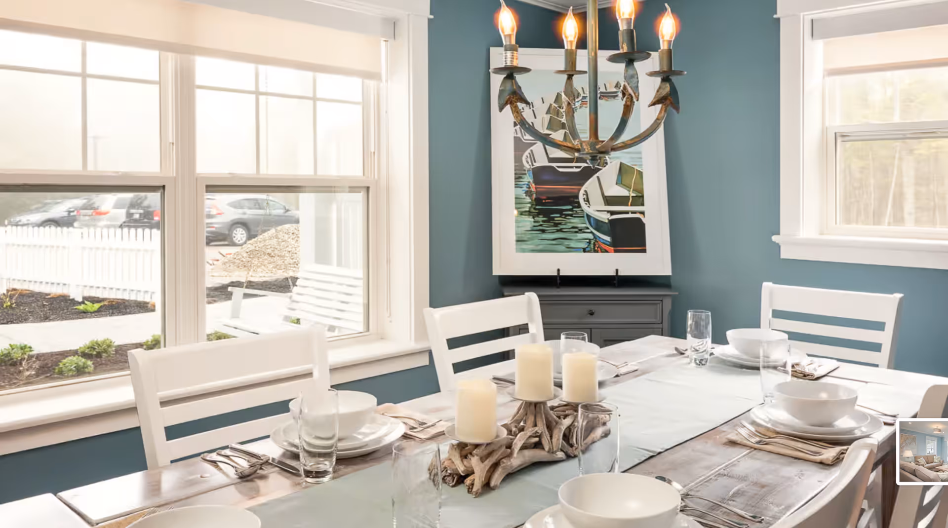Bright dining room with a rustic wooden table set for four, white chairs, a driftwood candle centerpiece, teal walls, a chandelier, and large windows.